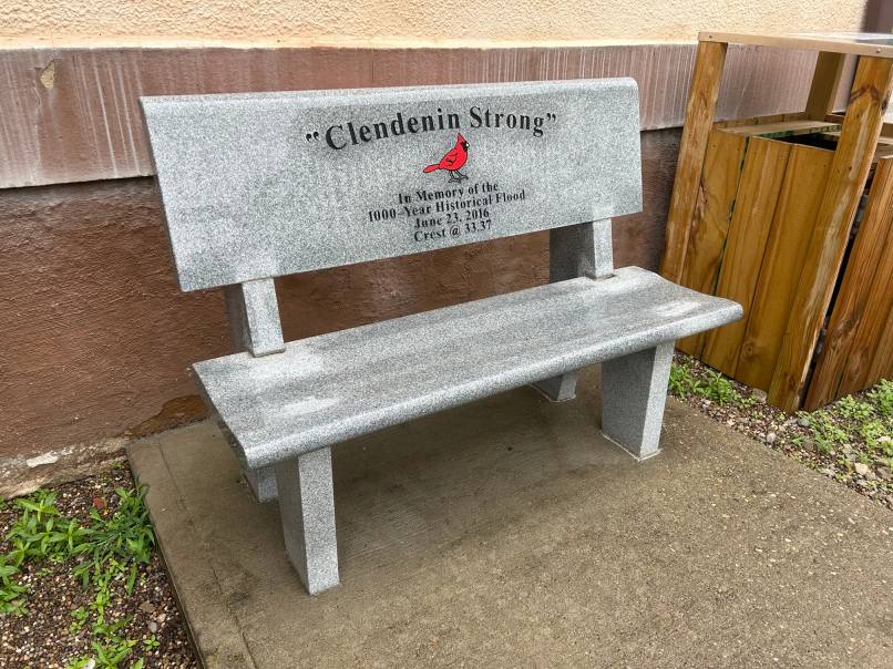 A bench engraved Clendenin Strong features a red cardinal and sits beside a building.